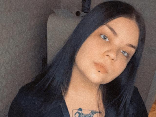 LottyAsya tiene el pelo negro, ojos azules, un piercing en la nariz, un piercing en el labio y un tatuaje en el pecho. Lleva una camiseta negra y está mirando ligeramente hacia un lado..