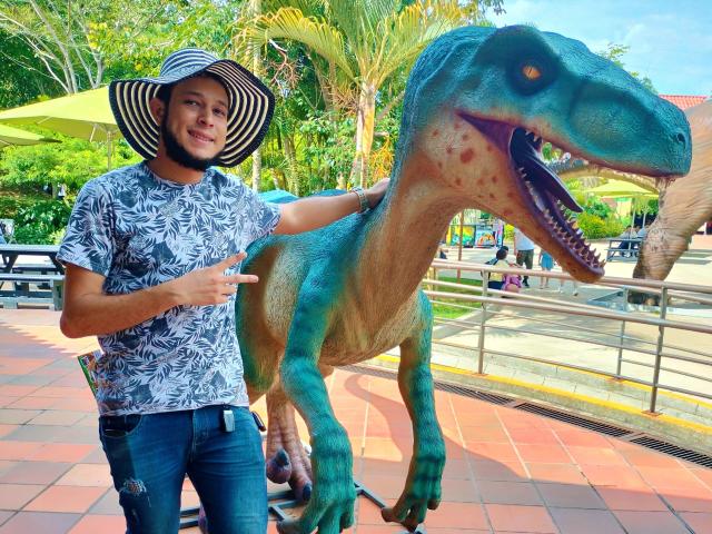 KalinDangerousCock with a wide-brimmed hat, wearing a floral shirt and jeans, standing next to a blue-green dinosaur statue, pointing with a smile. Dinosaur has a big open mouth.