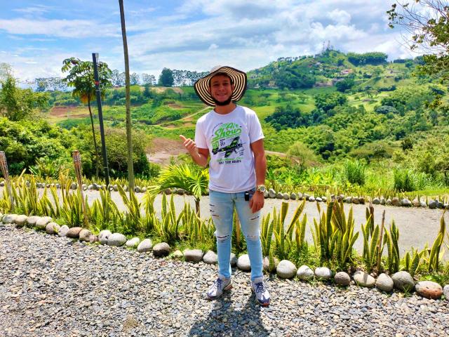 KalinDangerousCock está em frente a um caminho de cascalho, vestindo uma camiseta branca, jeans rasgados e um chapéu de palha, com um sorriso, apontando com um dedo e com uma montanha ao fundo..