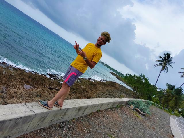 KalinDangerousCock está em uma beirada de concreto, vestindo uma camisa amarela, shorts roxos e rosa, e chinelos, com barba e cabelo cacheado, apontando com um sorriso..