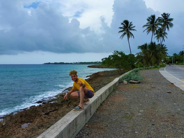 KalinDangerousCock, com cabelo loiro curto e encaracolado, camisa amarela e shorts roxos, está agachada em uma parede de concreto perto de uma costa rochosa com águas azuis, com palmeiras e uma estrada..