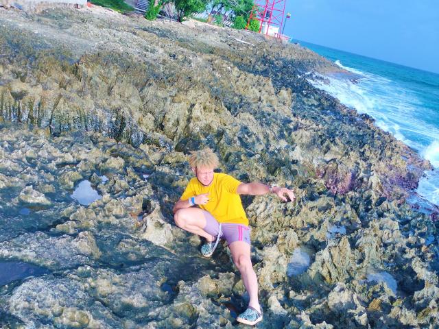 KalinDangerousCock, with curly blonde hair, wearing a yellow shirt and purple shorts, is crouching on rocky, wet shoreline, holding a small object, near crashing waves.
