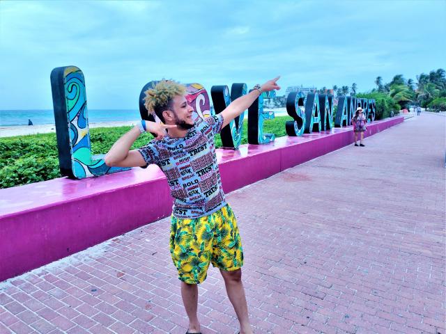KalinDangerousCock, com cabelos loiros encaracolados, vestindo uma camisa colorida com estampas e shorts florais amarelos, está descalça em um caminho de tijolos rosa, com os braços estendidos, em frente a letras grandes e azuis..