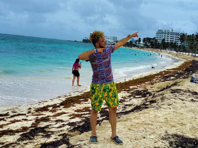 KalinDangerousCock, em pé em uma praia, vestindo uma camisa colorida e shorts com estampa verde e amarela, aponta com os braços estendidos, cabelo loiro, barba marrom, areia e algas sob os pés, oceano ao fundo..