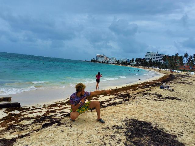 KalinDangerousCock with curly blonde hair, purple shirt, and green leaf in hand, crouches on sandy beach with seaweed, ocean in background, and overcast sky, standing woman in distance.