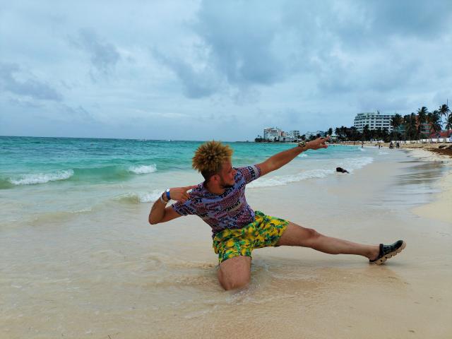 KalinDangerousCock, um homem loiro com barba, vestindo uma camisa colorida e shorts amarelo-esverdeados, está agachado em uma praia, alongando-se com um braço e uma perna..