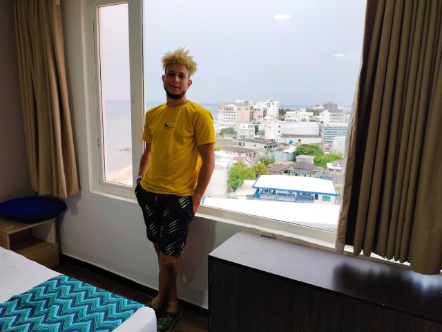 KalinDangerousCock está em um quarto vestindo uma camiseta amarela e shorts pretos com padrão verde, com as mãos nos bolsos, cabelo loiro curto e espetado, e um leve sorriso..