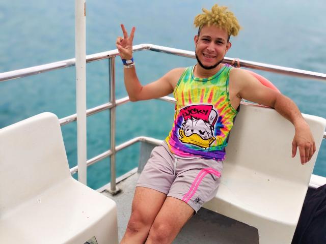 KalinDangerousCock, with curly blonde hair, a colorful tank top, and pink shorts, is sitting on a boat, flashing a peace sign, and smiling. KalinDangerousCock has a beard.