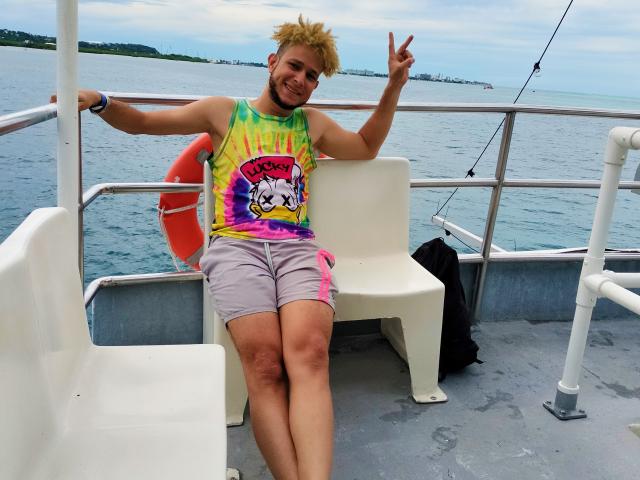 KalinDangerousCock, with spiky blonde hair, wearing a tie-dye shirt and pink-trimmed grey shorts, sits on a boat, flashing a peace sign, with a bruise on their thigh.