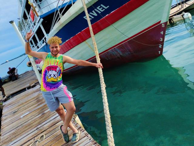KalinDangerousCock, com cabelo loiro, vestindo uma camisa verde neon e rosa, em pé em um cais, segurando uma corda, barco em água verde, camisa colorida com um desenho de dragão..