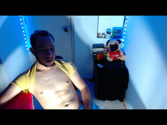 KalinDangerousCock com cabelo castanho curto, sem camisa, usando uma camisa amarela aberta, expondo um peito magro, tonificado, pálido e abdômen plano, sentado em um quarto com iluminação azul e segurando um brinquedo de pug..