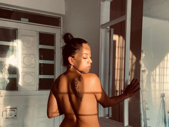 Amberv, topless, back to camera, brown skin, small feather tattoo on upper back, dark hair in bun, sunlight, door, shadow lines, right hand on door, eyes closed.