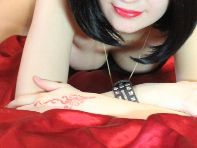 GloriaUpperLove, con cabello negro, lápiz labial rojo y un collar, inclinada, mostrando sus senos y mirando una mano con henna sobre seda roja..