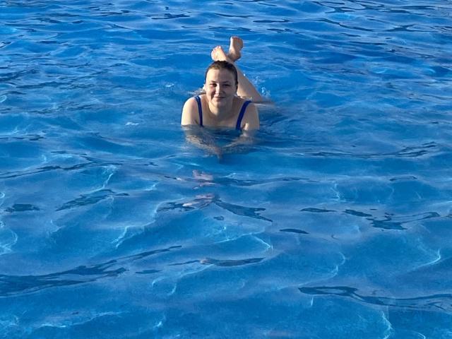 HanterHellen, les seins apparents, en maillot de bain bleu une pièce, allongée sur le ventre dans la piscine, les jambes levées, souriante, cheveux bruns, peau claire, l'eau reflétant une lumière bleue, surface de la piscine avec des ondulations..