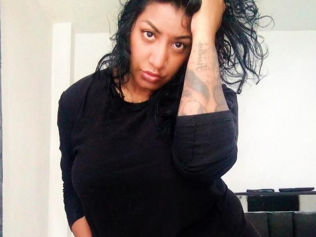 Nahirr with curly black hair, wearing a black long-sleeve shirt, looking at the camera with a slight pout, hand in hair, tattoo on right arm.