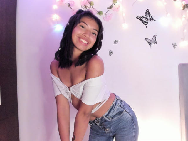 Lucia Garcia, met zwart haar, lachend, draagt een witte top met een open schouder die haar borsten onthult en blauwe jeans, leunt voorover en staat in een kamer met vlinderdecoraties aan de muur..