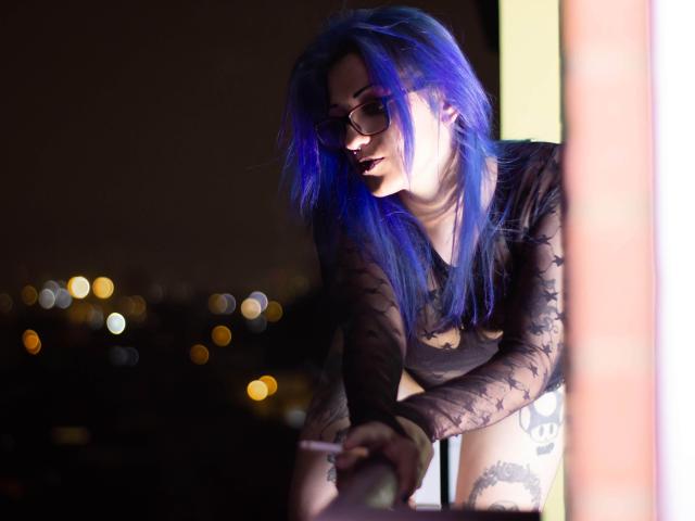 AnnyFreyr, avec ses cheveux bleus, ses lunettes et son haut transparent noir orné d'étoiles, penchée en avant, montrant sa cuisse tatouée, tenant une cigarette allumée par la lumière du soleil, avec un arrière-plan flou d'une ville nocturne..