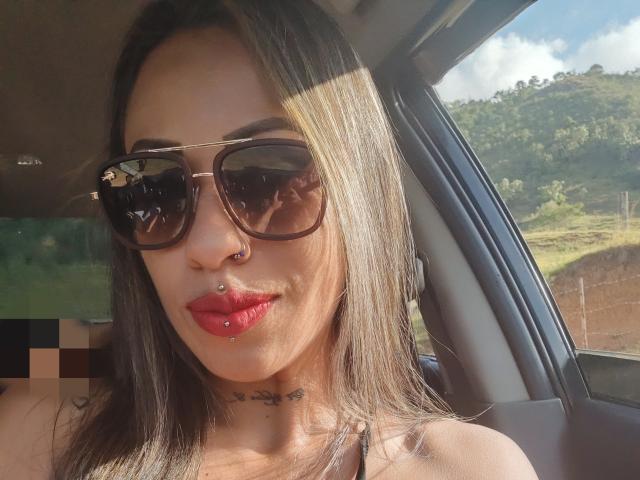LunaCandy, com cabelo castanho escuro, usando óculos de sol pretos grandes, batom vermelho, piercing no nariz, piercing no lábio e um colar, sentada em um carro. Seios não visíveis. Atrás não visível..