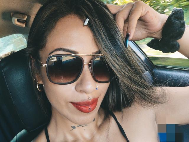 LunaCandy mit dunkelbraunen Haaren, goldenen Reifenohrringen, Nasenring, Septum-Piercing, rotem Lippenstift, Tattoo unter dem Kinn, schwarzem Haargummi, Sonnenbrille, blauen Nägeln, Autoinnenraum, Selfie..