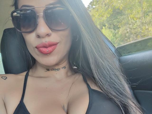 LunaCandy, com óculos de sol escuros, batom vermelho e um piercing no nariz, exibindo seus lábios com piercings e um sutiã preto, sentada em um carro com seus seios e tatuagens à mostra..