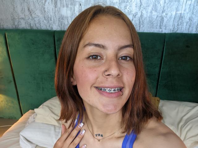 VictoriaOrtega with brown hair, nose ring, and blue tank top, smiling with metal braces on her teeth, hand on her chest, and "TBA" tattoo below her collarbone.