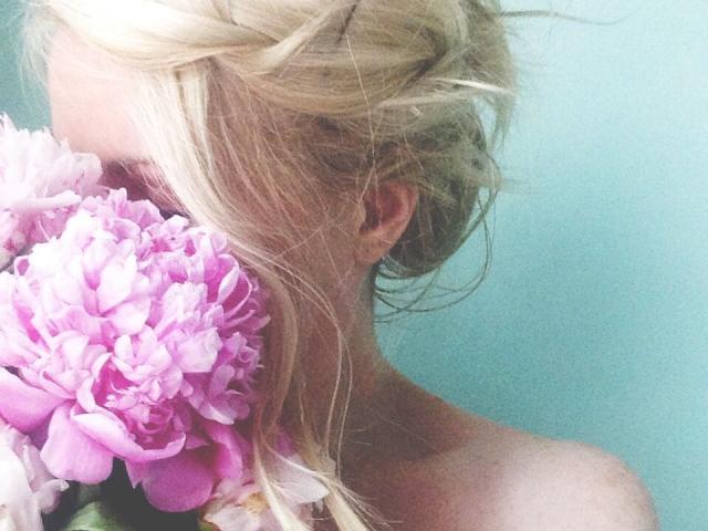 I capelli biondi di LambaKaif sono raccolti in un disordinato chignon, e tiene dei fiori di peonia rosa vicino al viso, coprendo il seno. Le sue spalle nude sono visibili su uno sfondo verde tenue..