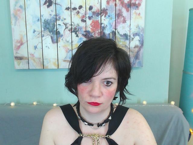 EverlyRays has short black hair, red lipstick, and black harness with metal rings, looking at the camera with a slight blush on her pale skin, and a serious expression.
