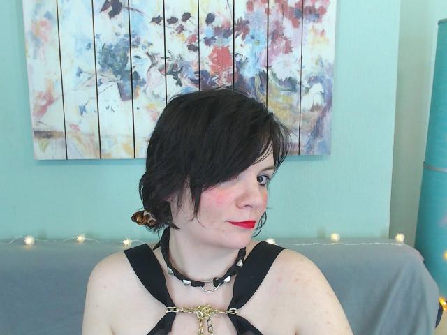 EverlyRays with short black hair, red lipstick, black harness, spiked choker, looking to the side, fair skin, light blush, small breasts, sitting in front of a colorful abstract painting.