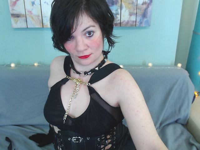 EverlyRays with short black hair, red lipstick, and pale skin, wearing a black harness with gold chain, showing her tits, sitting on a grey-covered couch.