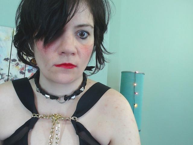 EverlyRays with short black hair, red lipstick, black harness, chain, and spiked collar, looking at camera, pale skin, small tits, slight blush on cheeks, green wall, blue lamp.