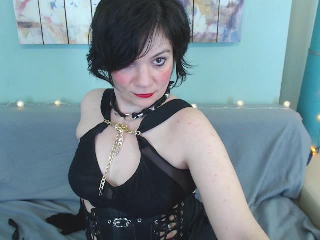 EverlyRays with short black hair, red lipstick, black sheer top with chain, and corset, showing off her tits, wearing a choker, sitting on a grey bed.