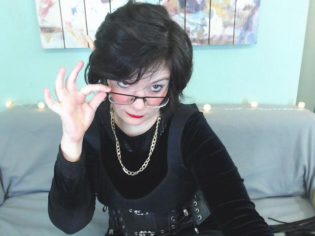 EverlyRays with short black hair, red lipstick, black dress, gold chain, and glasses, adjusting them with her right hand, sitting on a gray couch.