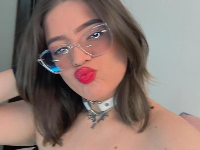 Kim, ein Sommerbild mit braunen Haaren, trägt eine klare Brille und eine weiße Krawatte mit einem Metallkreuz, hat leicht gerötete Lippen, und in der Brille spiegelt sich Blau, wobei nackte Schultern zu sehen sind..