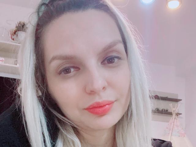 DouceAmelia has light skin, blue eyes, and red lipstick. She's smiling with blonde and dark brown hair, wearing a black top. Her tits are not visible.