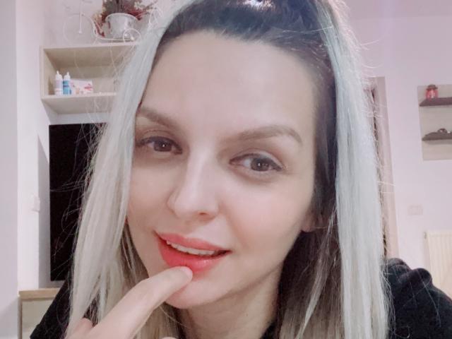 DouceAmelia with light blonde hair, brown eyes, and red lipstick, touching her red lips with her index finger, smiling gently. She's wearing a black top.