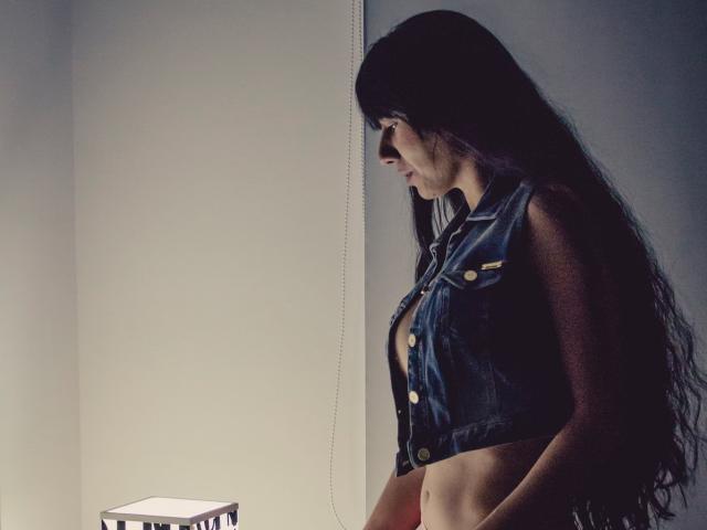 XiaQueen, avec ses longs cheveux noirs, portant un gilet en jean sans manches, exposant sa poitrine nue, se tient de profil et regarde une boîte allumée posée sur une table..