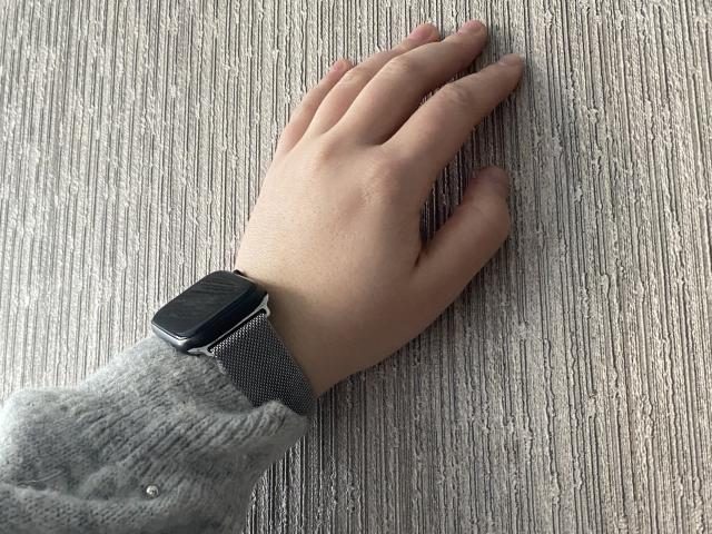 DiLaria's hand with a gray wristwatch, wearing a fuzzy gray sleeve, resting on a textured wooden surface. DiLaria's fingers are slightly spread.