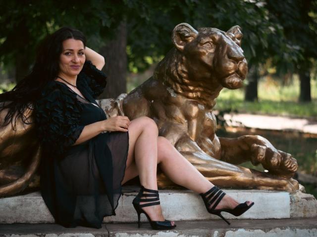 GinaONeon sits on a bronze lion statue, wearing a black sheer dress and high heels, with long dark hair and a slight smile, showing her ass and legs.
