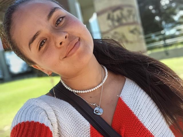 Arielle Wilde hat ein süßes Gesicht, lächelt leicht mit braunen Augen, trägt einen roten und weißen Strickpullover, eine Perlenkette und einen schwarzen Träger, und hat braunes, langes Haar zu einem Pferdeschwanz gebunden..