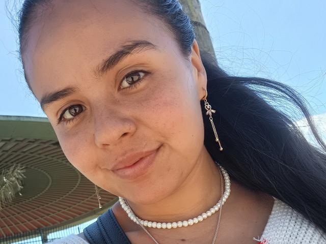ArielleWilde has tan skin, dark eyebrows, and long black hair. She's smiling with a pearl necklace, earrings, and a white top. Her brown eyes are looking at the camera.