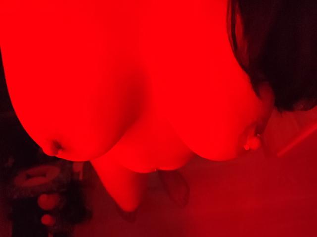 StefyLoveSex' große, rot getönte Brüste hängen herab, wobei ihre Brustwarzen leicht sichtbar sind, und ihr dunkles Haar ist teilweise im Bild zu sehen, beleuchtet von rotem Licht..