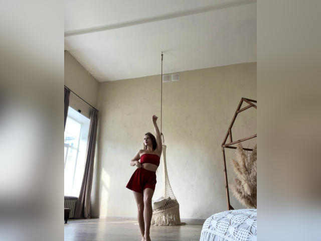 Peony, con un top y pantalones cortos rojos, levantando el brazo, de pie frente a una silla colgante, suelo de madera, paredes beige, luz del sol entrando por la ventana, mostrando su trasero desnudo y pequeños pechos..