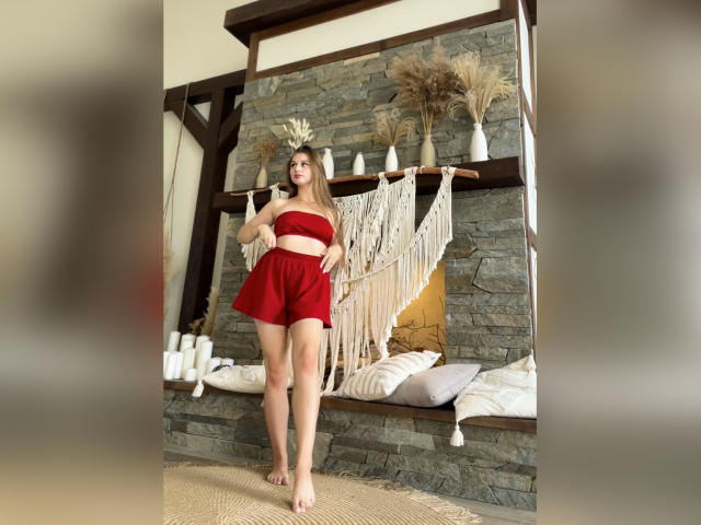 PeonyAmazing staat blootsvoets in een rode top en korte broek, met haar top in haar handen, met lang blond haar, voor een stenen openhaard, een macramé wanddecoratie en witte kaarsen..