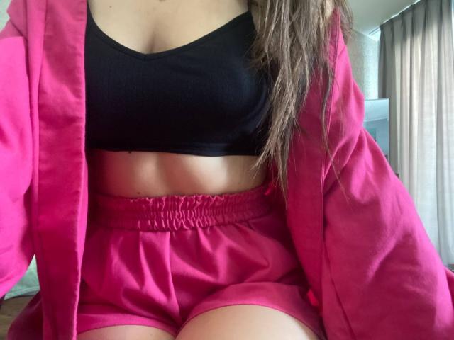 CheriLady, avec un short rose, un débardeur de sport noir et une veste rose, montrant son ventre et ses petites seins, ses cheveux bruns et un léger bronzage, assise sur un lit..
