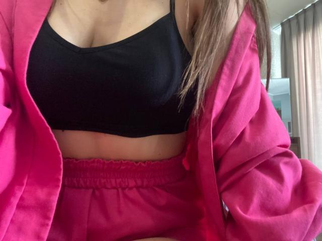 CheriLady portant une veste rose par-dessus un soutien-gorge de sport noir et un short rose, montrant ses seins et une partie de son ventre plat. Ses cheveux bruns sont partiellement visibles..