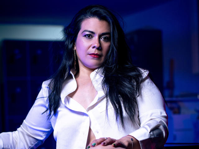 MartinaFox69 has long black hair, red lipstick, and a white button-up shirt, slightly open, with her tits partially visible, sitting with a serious, slightly annoyed expression.