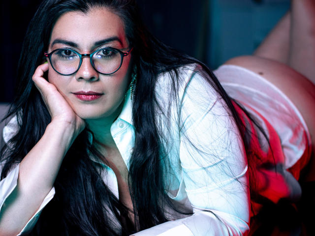 MartinaFox69 with long black hair, glasses, white shirt, and red lighting, lying on her stomach, butt up, hand on cheek, looking at camera, serious expression, sexy.