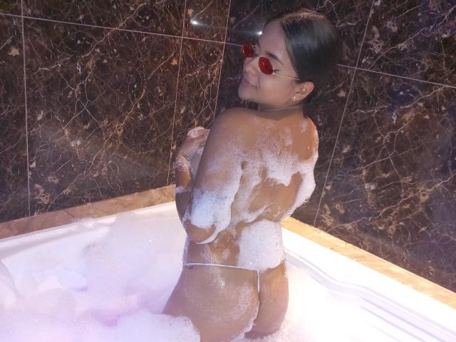 MeganBrownish in a bathtub, covered in bubbles, wearing red sunglasses, looking back, showing her ass, with soap suds on her back and butt, dark brown marble tiles.