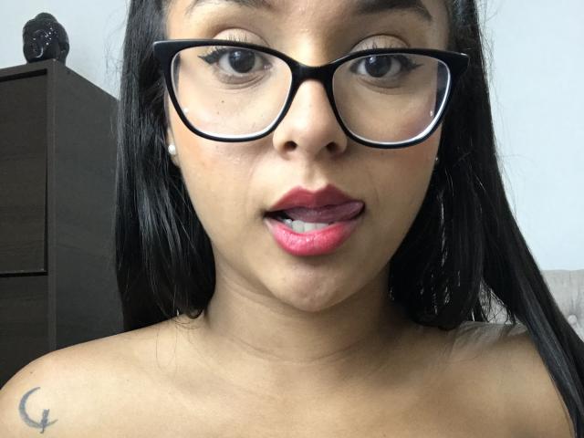 Megan Brownish, con gafas oscuras, lápiz labial rosa y la lengua afuera, mostrando su piel morena y cabello negro, tiene un pequeño tatuaje en forma de media luna en el hombro izquierdo..