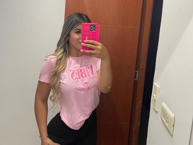 SashaPolet with long blonde hair, pink shirt, black skirt, holding pink phone, taking mirror selfie, showing her tits, in a room with wooden door and white walls.